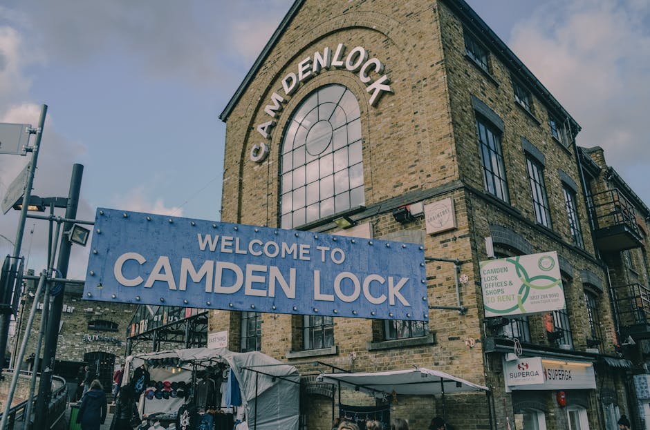 A busy street scene in Camden, Hampstead, featuring a large railway bridge with the word 'Camden Lock' painted in bold yellow and orange letters on a green background. The bridge is supported by metal