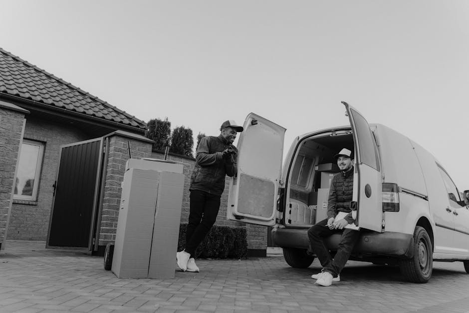 Two young women sitting inside the open rear of a moving van engaged in conversation, with one of them gesturing with her hand while the other smiles. The interior of the van shows several cardboard b
