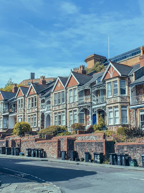 A busy street scene in Hampstead with multiple multi-storey buildings lining the road, some with flower baskets hanging from their facades. The buildings feature a mix of brick and painted exteriors,